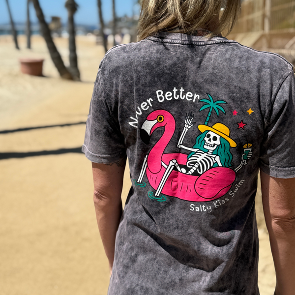 Person wearing a t-shirt with a flamingo and skeleton design on a beach.