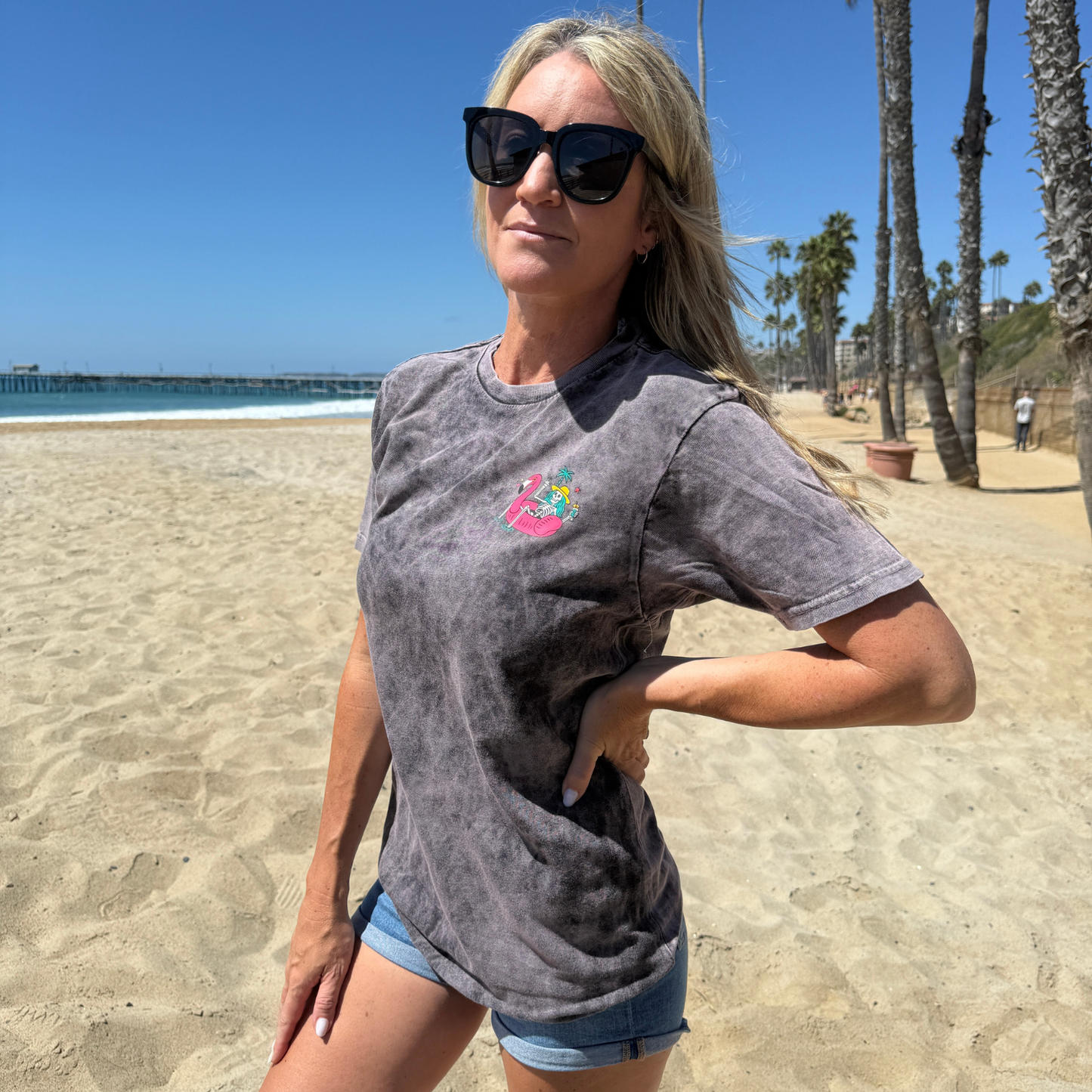 Woman wearing sunglasses and a gray t-shirt with a colorful design on a beach.