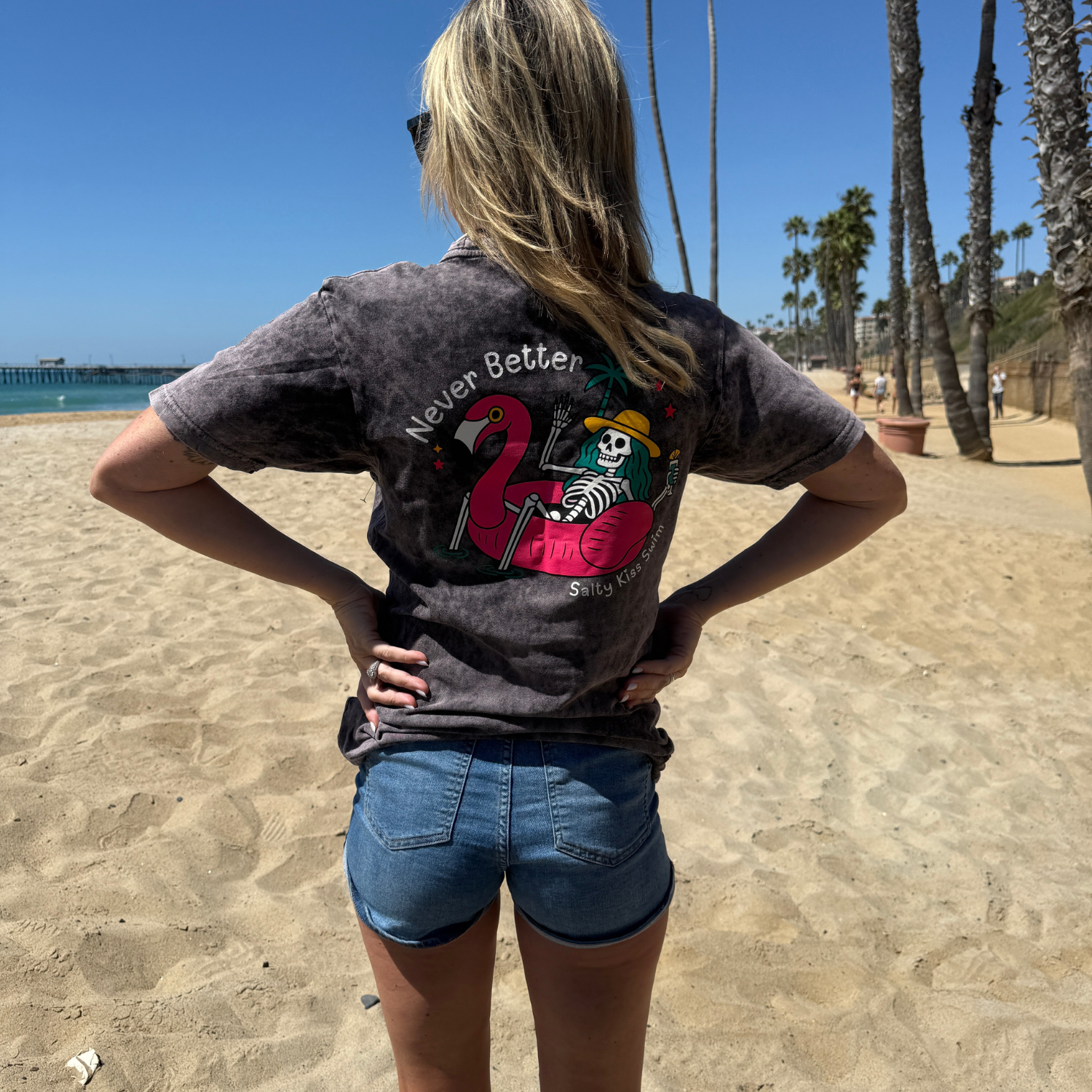 Person wearing a t-shirt with a flamingo and skeleton design on a beach.