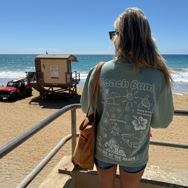 Person wearing a 'Beach Bum' shirt on a beach with ocean and lifeguard stand in the background.