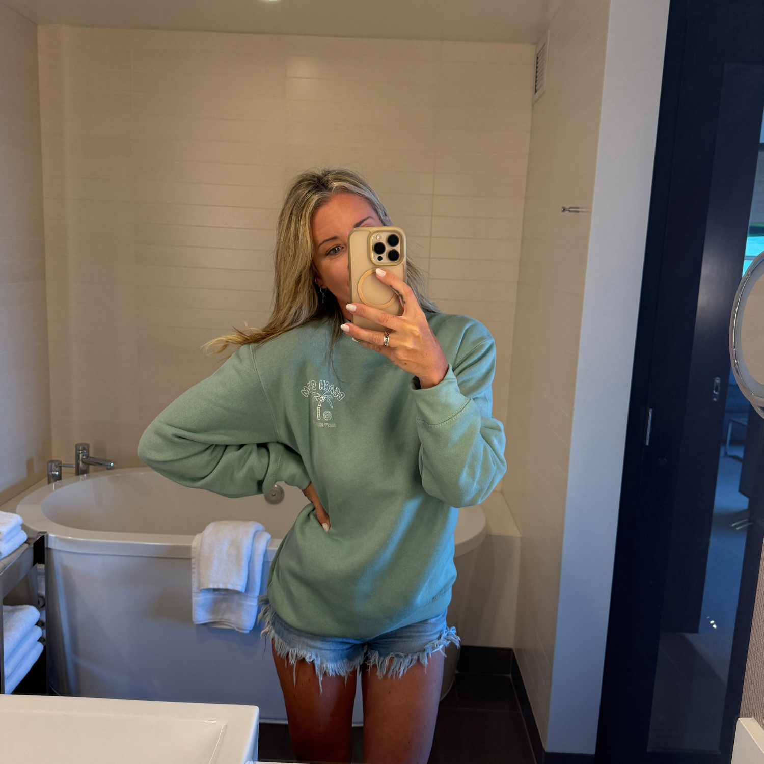 Person taking a mirror selfie wearing a light green sweatshirt and denim shorts in a bathroom.