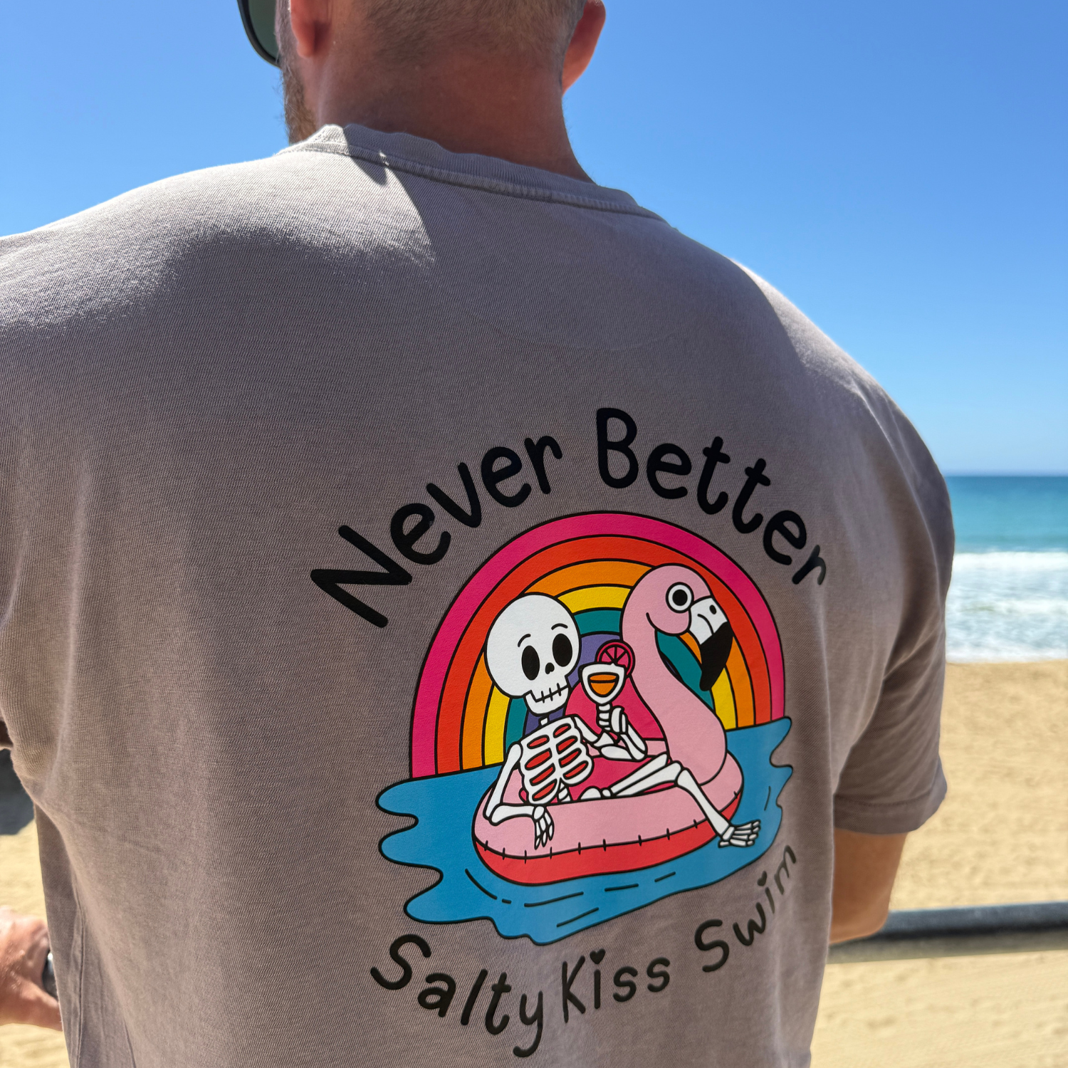 Person wearing a t-shirt with a skeleton and flamingo design and text 'Never Better Salty Kiss Swim' at the beach.