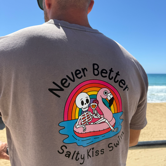 Person wearing a t-shirt with a skeleton and flamingo design and text 'Never Better Salty Kiss Swim' at the beach.